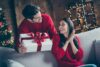karácsonyi ajándékötletek nőknek – egy férfi megajándékoz egy nőt, mindketten piros pulóverben vannak, ünnepi hangulat leli őket körbe