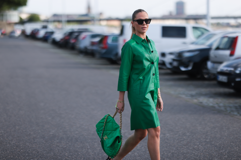 Nő egy zöld egyberuhában, és láncos táskával sétál a parkolóban