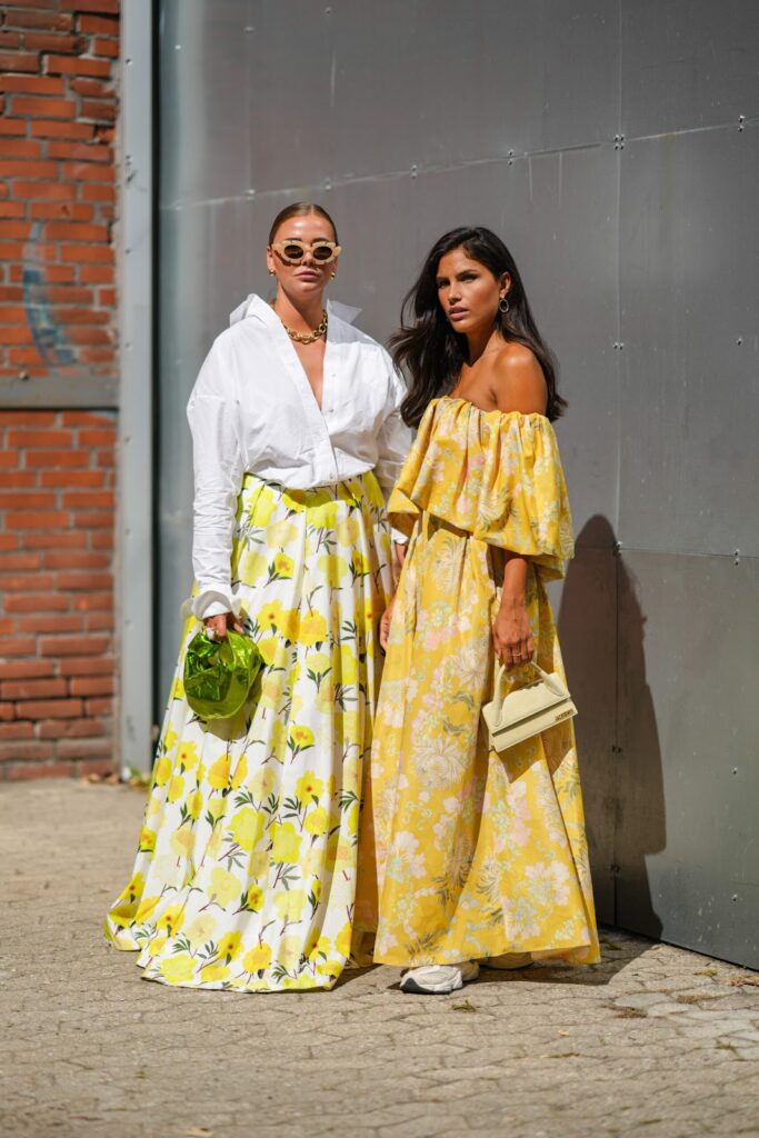 Tavaszias ruhába öltözött nők – egy fehér túlméretezett ing egy virágos maxiszoknyába bújtatva, valamint egy sárga virágmintás ruha és tornacipő.